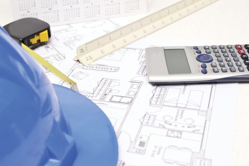 Close-up of architectural blueprints with a blue hard hat, calculator, ruler, and measuring tape.